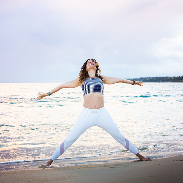 Beautiful woman in white leggings and stripe shirt stands in a power pose with open arms rising to the sky as the sun is rising in the background at this epic Maui beach.