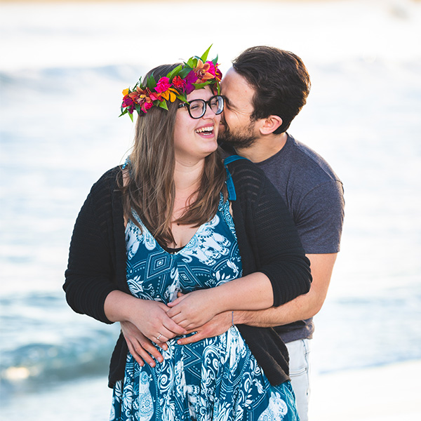 Happy couple dressed in blues and greens with a flower head lei are holding each other laughing at the beach during this sunrise Maui couple photography session.