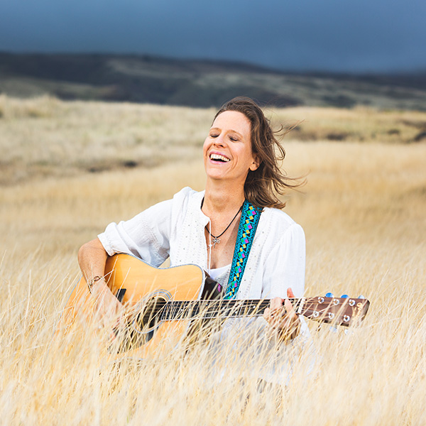 Maui portrait photography session of a beautiful woman in white blouse plays guitar and sings in golden grass with a rich dark blue sky in the background.