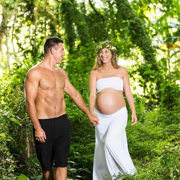 Maui maternity photography session of a shirtless muscular man holding the hand of his beautiful pregnant wife as they smile and happily walk through the bright green forest.